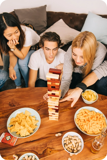 Drie vrienden spelen een spelletje Jenga aan een houten tafel, omringd door snacks zoals chips en pinda's. Ze lijken geconcentreerd en betrokken.